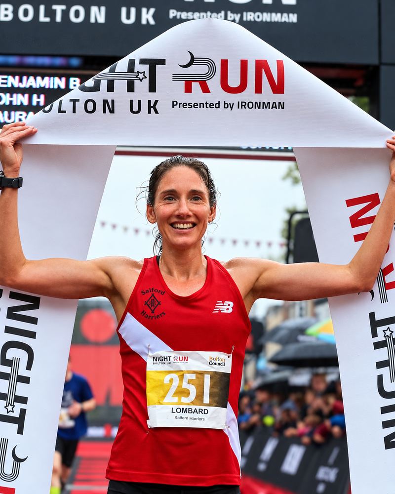 Laura Lombard smiling as she holds the finish tape after winning the Night Run Bolton 5K, wearing a red Salford Harriers vest